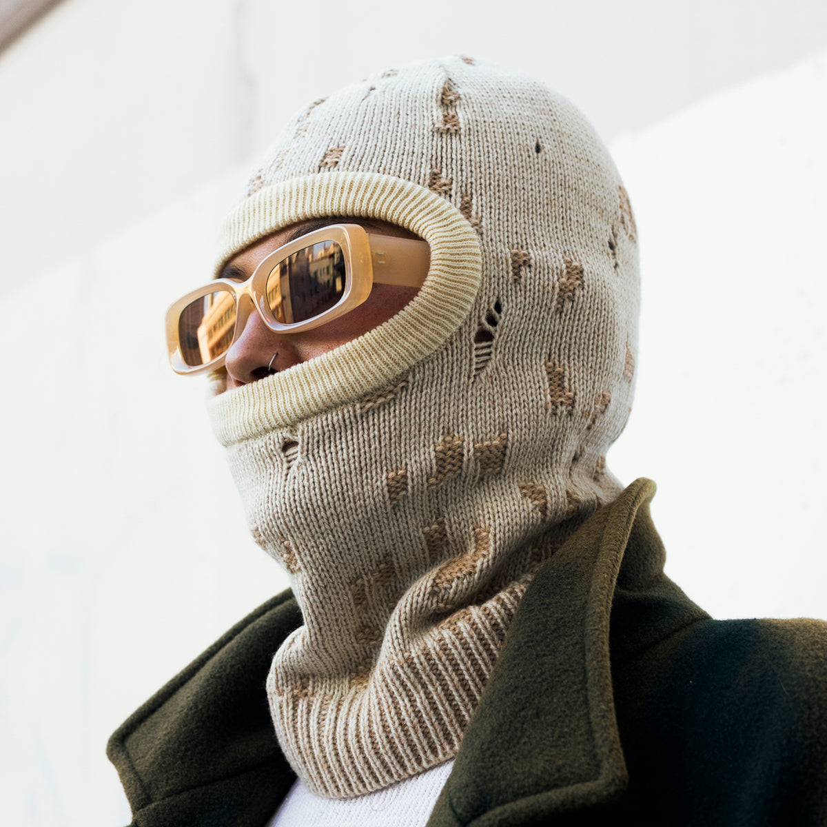A person wearing a beige knit balaclava mask with distressed details, gold-framed sunglasses, and a dark olive green coat against a bright white background.